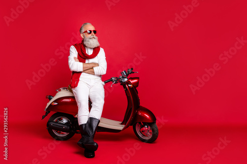 Aged macho man sitting on retro moped with crossed arms wear jumper and trousers isolated red background