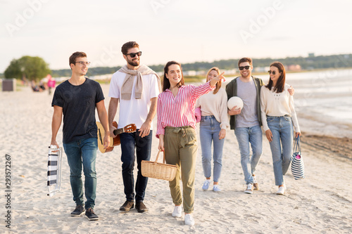 friendship, leisure and people concept - group of happy friends with guitar, chair and picnic basket walking along beach in summer