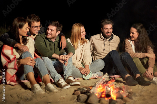 leisure and people concept - group of smiling friends sitting at camp fire on beach at night