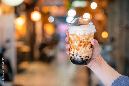 Young woman is holding, drinking brown sugar flavored tapioca pearl bubble milk tea with glass straw in night market of Taiwan, close up, bokeh