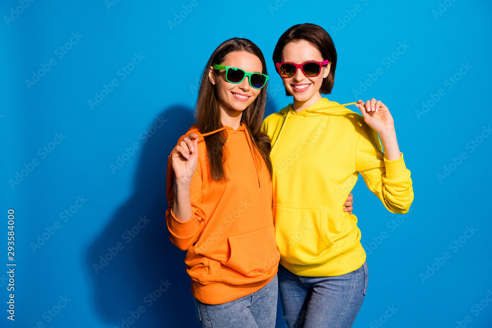 Photo of pretty two girlfriends lady standing hugging ready for holidays and rest wear sun specs casual bright hoodies and jeans isolated blue color background Photo of pretty two girlfriends lady standing hugging ready for holidays and rest wear sun specs casual bright hoodies and jeans isolated blue color background