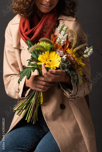 cropped view of woman in trench coat holding bouquet of autumnal wildflowers on black background