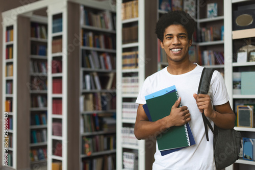 Fotografie, Obraz Handsome afro student posing on bookshelves background