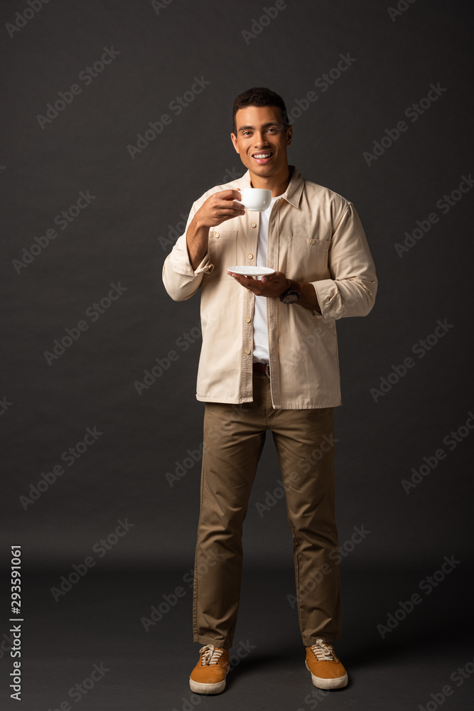 smiling handsome mixed race man in beige shirt holding cup of coffee and saucer on black background