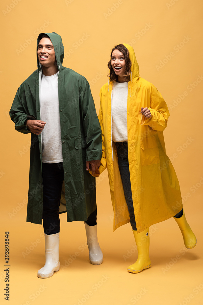 happy interracial couple in raincoats and rubber boots on yellow background