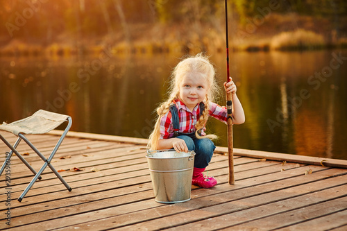 Little girl fisherman with a fishing rod and a bucket of fish on a wooden pier.