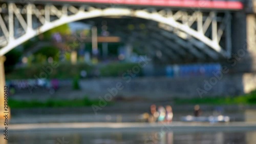 Blurred background of bridge across the river, with people sitting on the waterfront on a warm summer evening. photo