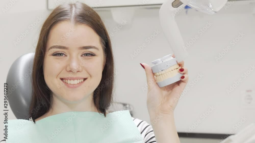 Beautiful cheerful woman smiling holding teeth model. Attractive female patient holding jaw mold waiting for dental checkup at the clinic. Health, medicine concept Beautiful cheerful woman smiling holding teeth model. Attractive female patient holding jaw mold waiting for dental checkup at the clinic. Health, medicine concept