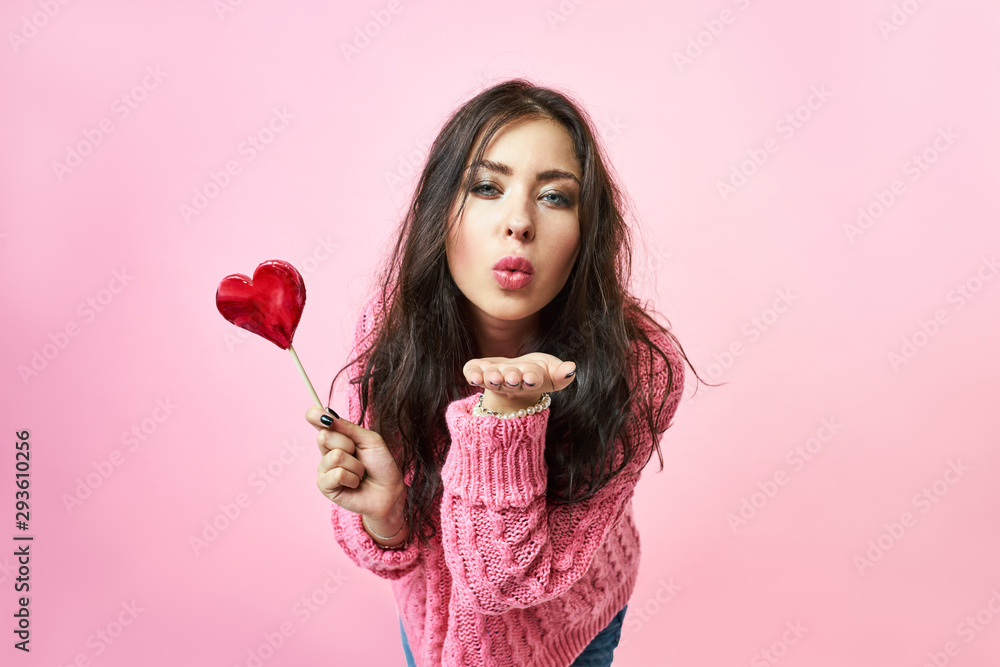 Portrait of a cute girl with a heart shaped lollipop. Sends blows a kiss while looking at the camera, Sexy female lips. Separate on a white background. Portrait of a cute girl with a heart shaped lollipop. Sends blows a kiss while looking at the camera, Sexy female lips. Separate on a white background.