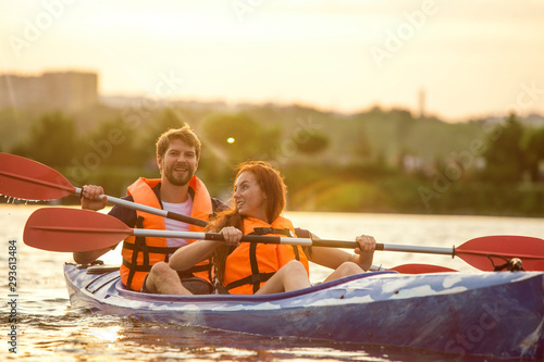 Happy young caucasian couple kayaking on river with sunset in the backgrounds. Having fun in leisure activity. Happy male and female model laughting on the kayak. Sport  relations concept. Colorful.