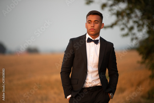 A handsome teenager dressed in black suit and bow tie and looks serious, He stands on a field that has recently been harvested in the sunset 