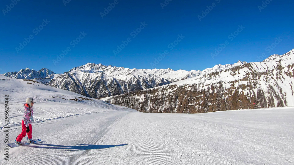 A snowboarder going down the slope in Heiligenblut, Austria. Perfectly groomed slopes. High mountains surrounding the girl, wearing pink trousers and colorful jacket. Girl wears helm for protection