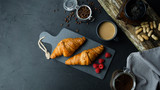 Rogale croissant leżące na ciemnej desce w otoczeniu malin i kawy. Ciemne tło, widok z góry.