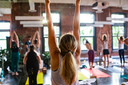Diverse group of people in yoga class. A young Caucasian girl with blonde hair tied in a pony tail is seen from the rear as she does a standing pose during 108 rounds of surya namaskar.