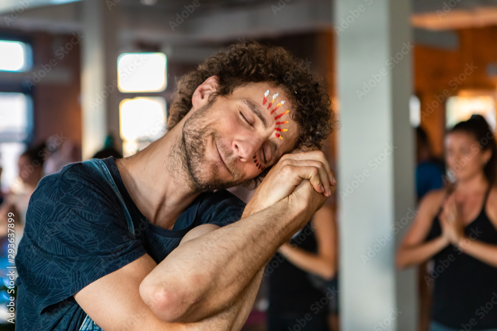 Diverse group of people in yoga class. A young and spiritual Caucasian guy is seen up close, resting head on clasped hands with locked arms. Relaxed pose during 108 salutations to the sun. Diverse group of people in yoga class. A young and spiritual Caucasian guy is seen up close, resting head on clasped hands with locked arms. Relaxed pose during 108 salutations to the sun.
