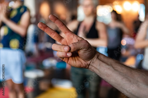 Diverse group of people in yoga class. The hand of a motivational guy is viewed closeup, with fingers giving the v sign inside a gym as blurry people are seen in yogic pose in background.