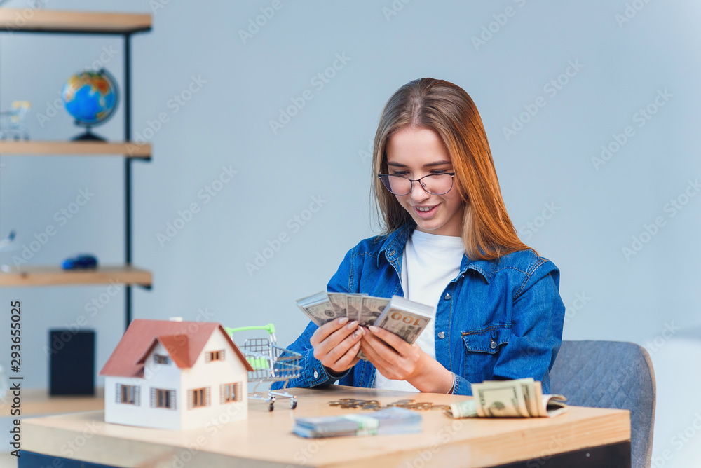 Young pretty girl counts her savings money and looks to a model of the house. Young pretty girl counts her savings money and looks to a model of the house.