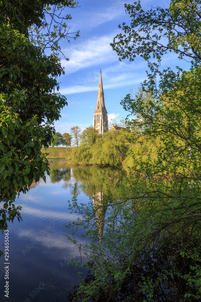 Tower of St. Alban's (English) church in Copenhagen, Denmark. Blue sky and green trees and vegetation are reflected in water in the foreground. Sunny afternoon in spring.