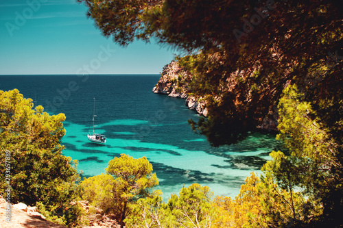 Lovely and beautiful empty beach view from above with a sailing boat in the turquoise ocean sea water and calm and soft summer morning light framed by pine tress. Beach, Mallorca, Spain