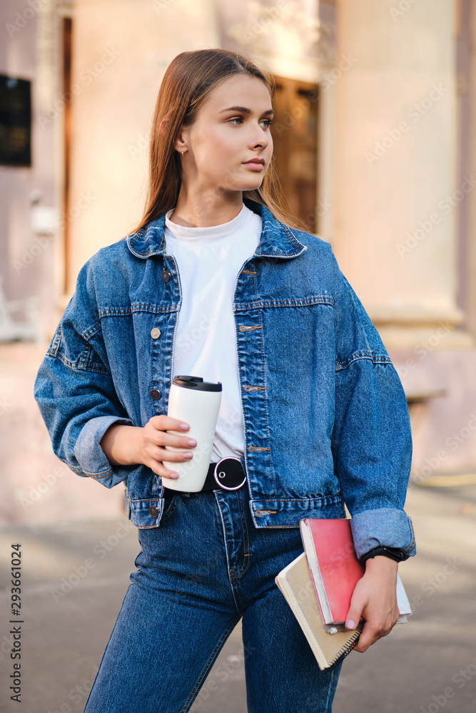 Attractive stylish student girl in denim jacket with coffee and textbooks thoughtfully looking away outdoor Stock Adobe Stock