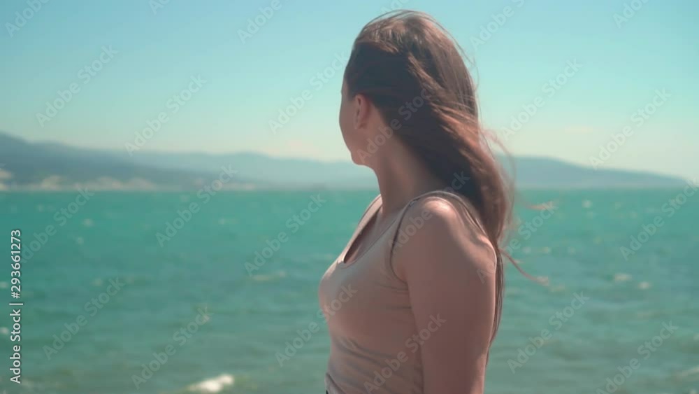 A girl in a T-shirt with long hair is sitting near the sea. A girl sits on the embankment against the backdrop of mountains, sky and sea, her hair develops in the wind.