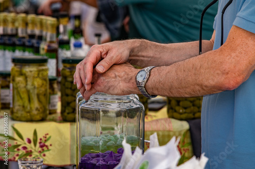 Fresh food at outdoor farmer's market. A closeup view on the arms and hands of an elderly Caucasian male wearing a wrist watch, standing by glass jars of foodstuff on a market stall