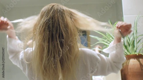 A blonde women doing her hair in front of the bathroom mirror in a dressing gown, shot from behind 4K