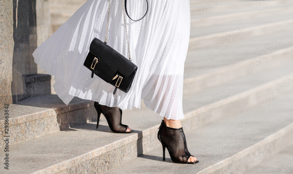Fashionable bag close-up in female hands.Girl walks in the city outdoors. Stylish modern and feminine image, style. Girl in a white light dress with a black bag and black boots, or heeled shoes. - obrazy, fototapety, plakaty 