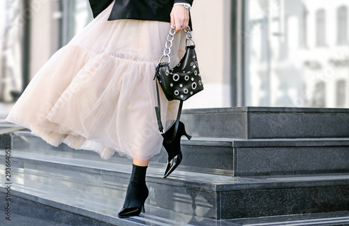 Fashionable bag close-up in female hands.Girl walks in the city outdoors. Stylish modern and feminine image, style. Girl in a white light puffy dress with a black bag, black boots or high-heeled shoes