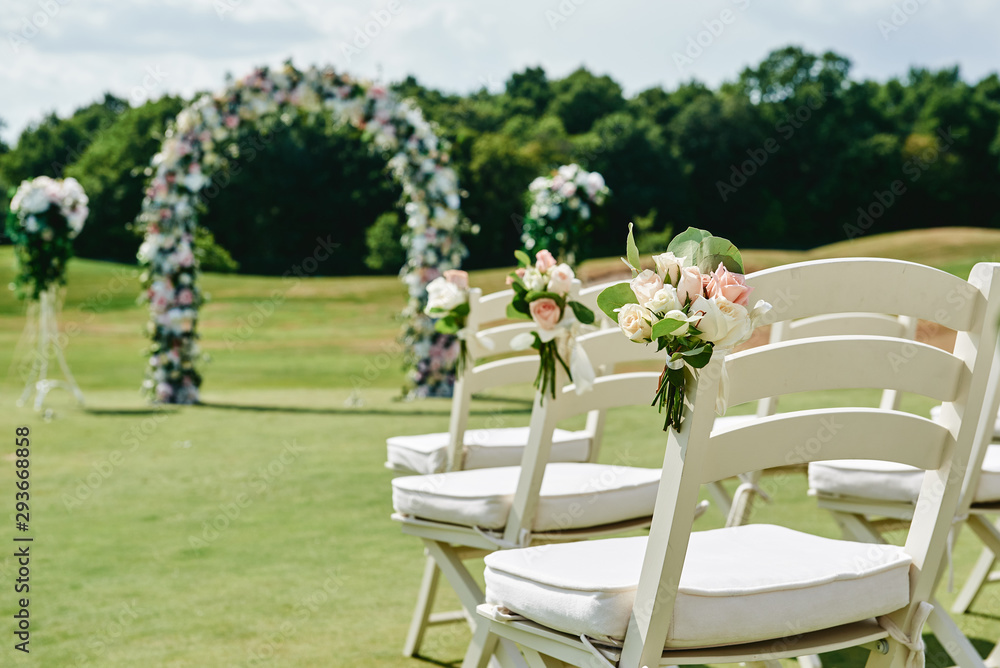 White wooden chairs with rose flowers on each side of archway outdoors, copy space. Empty chairs for guests prepared for wedding ceremony on golf course White wooden chairs with rose flowers on each side of archway outdoors, copy space. Empty chairs for guests prepared for wedding ceremony on golf course