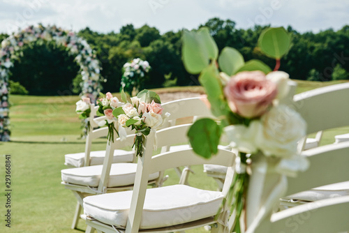 White wooden chairs with rose flowers on each side of archway outdoors, copy space. Empty chairs for guests prepared for wedding ceremony on golf course