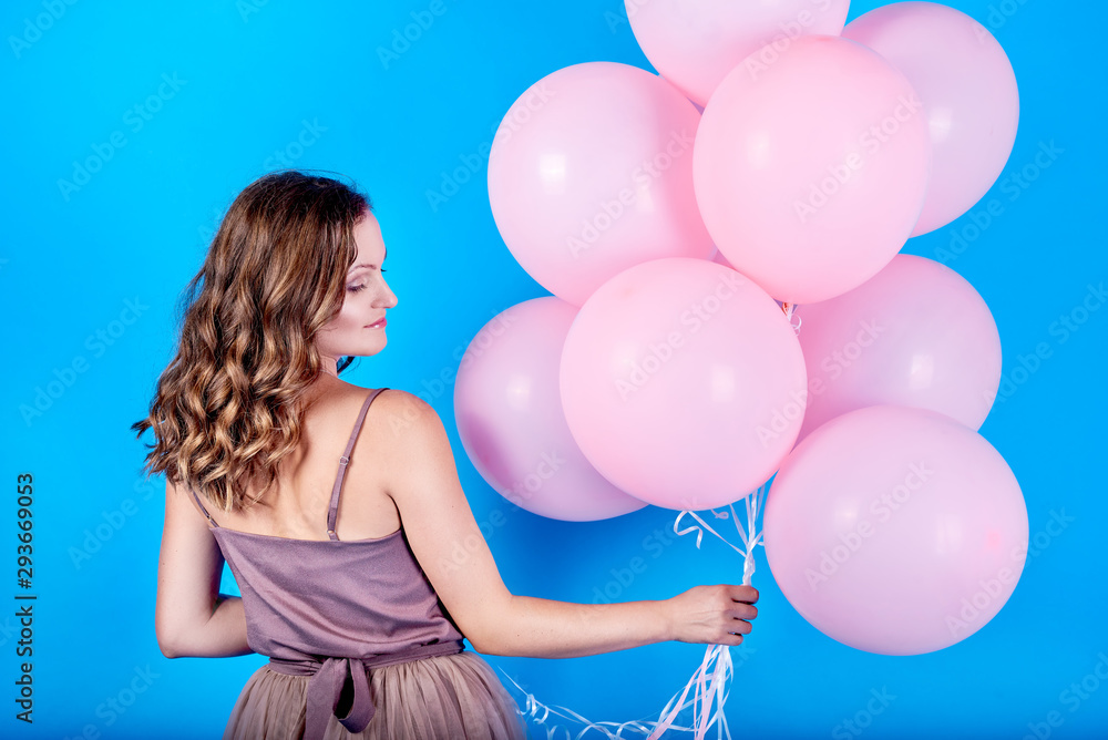 Obraz premium Beautiful young woman in dress holding pink balloons over blue background, back view. Holiday, birthday, valentine, fashion concept