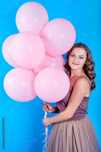 Happy smiling young woman in dress holding pink helium air balloons over blue background. Holiday, birthday, valentine, fashion concept