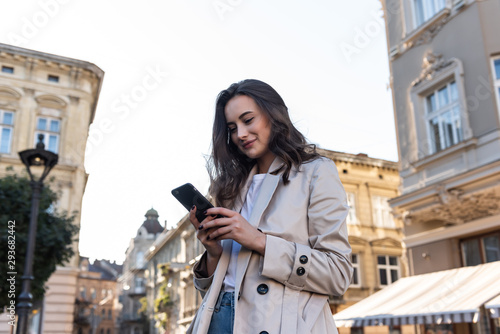 Adult caucasian confident young businesswoman is using smartphone app outside near modern office building.