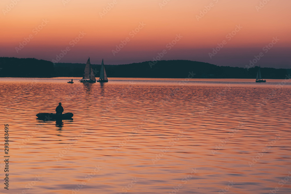 man fishing on a lake from the boat at sunset. silhouette of fishermen. yellow and orange sunset background. sailing boats in water at evening.