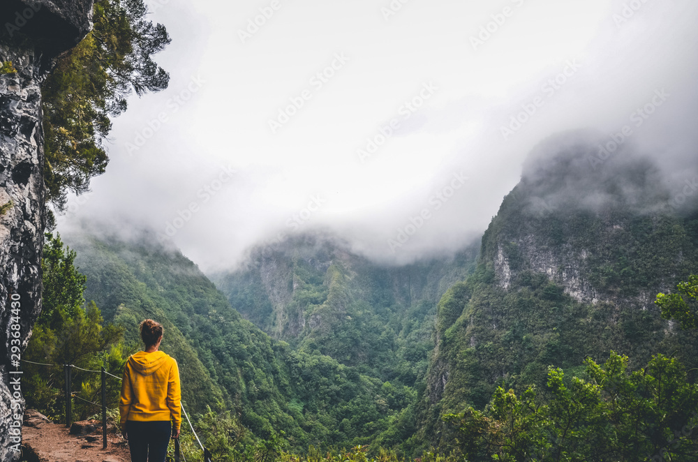 Young woman in yellow sweatshirt on a viewpoint in Levada Caldeirao Verde, Madeira, Portugal. Green scenic mountains in fog, misty landscape. Female traveler. Instagram, hipster filter