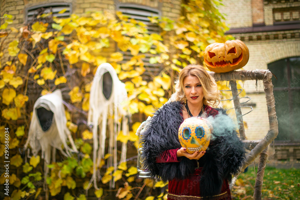 Beautiful woman in a witch costume holds a pumpkin painted in ack-o-lanterns style. Steaming pumpkin in the decor for Halloween. Sorceress on a background of ghosts.