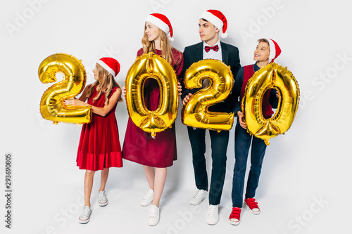 Young happy family with kids wearing Santa hats holding balloons shaped like numbers 2020 on white background.