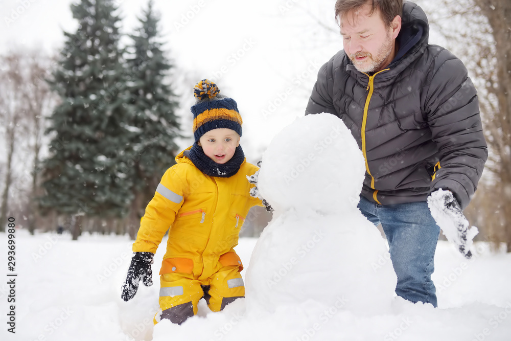 Little boy with his father building snowman in snowy park. Active outdoors leisure with children in winter.