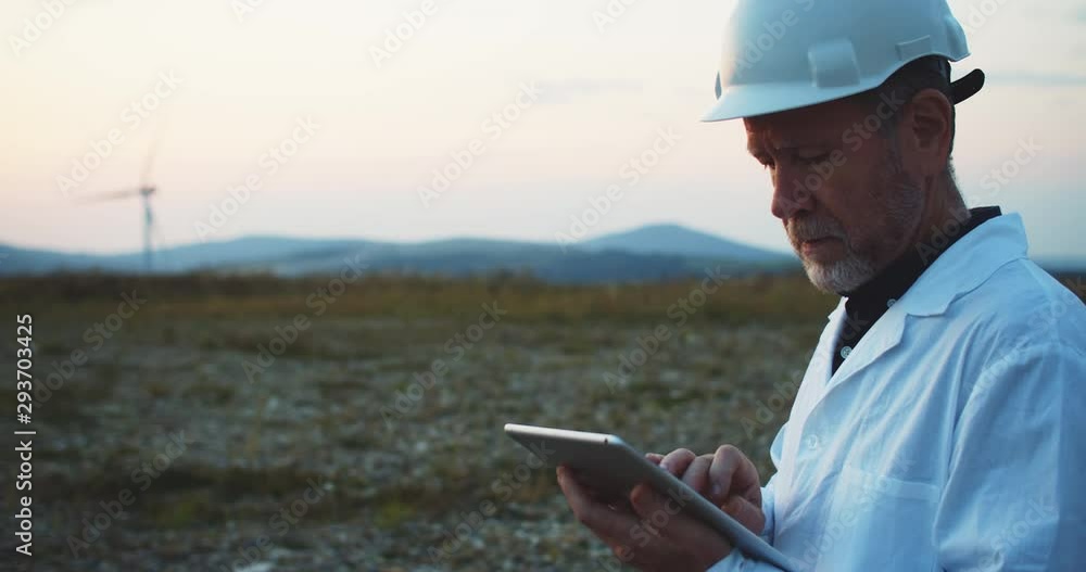 Experienced male technician in hardhat browsing app on digital tablet using modern technology while working at wind generating power station. Experienced male technician in hardhat browsing app on digital tablet using modern technology while working at wind generating power station.