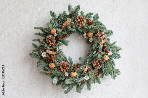 Christmas wreath made of natural fir branches  hanging on a white wall.  Wreath with natural ornaments: bumps, walnuts, cinnamon, cones. New year and winter holidays. Christmas decor