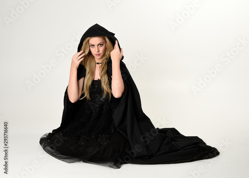 full length portrait of blonde girl wearing long black flowing cloak, sitting on the floor  with  a white studio background.