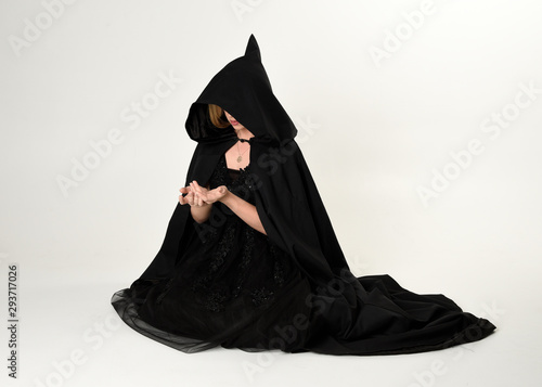 full length portrait of blonde girl wearing long black flowing cloak, sitting on the floor  with  a white studio background.