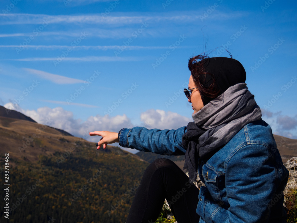 The girl shows a finger to the distance towards the mountains. A girl in a denim jacket, sunglasses and a scarf on her head The girl shows a finger to the distance towards the mountains. A girl in a denim jacket, sunglasses and a scarf on her head