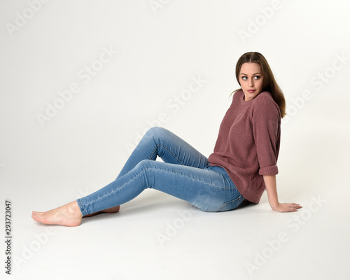 full length portrait of brunette woman wearing jeans and pink jumper. seated pose with a white studio background.