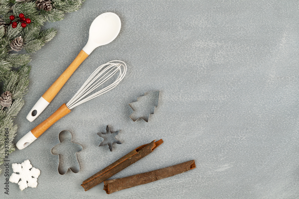 Utensils for Christmas baking with spoon, whisk, cookie cutters, cinnamon sticks and snow flake with snowy pine branches with cones over stone like background with copy space. Flat lay, top view. Utensils for Christmas baking with spoon, whisk, cookie cutters, cinnamon sticks and snow flake with snowy pine branches with cones over stone like background with copy space. Flat lay, top view.