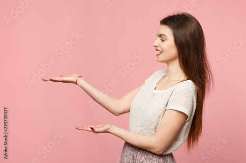 Side view of smiling young woman in casual light clothes posing isolated on pink wall background, studio portrait. People sincere emotions lifestyle concept. Mock up copy space. Pointing hands aside. © ViDi Studio