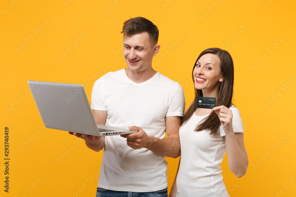Smiling young couple two friends guy girl in white t-shirts posing isolated on yellow orange background. People lifestyle concept. Mock up copy space. Holding laptop pc computer, credit bank card. Smiling young couple two friends guy girl in white t-shirts posing isolated on yellow orange background. People lifestyle concept. Mock up copy space. Holding laptop pc computer, credit bank card.