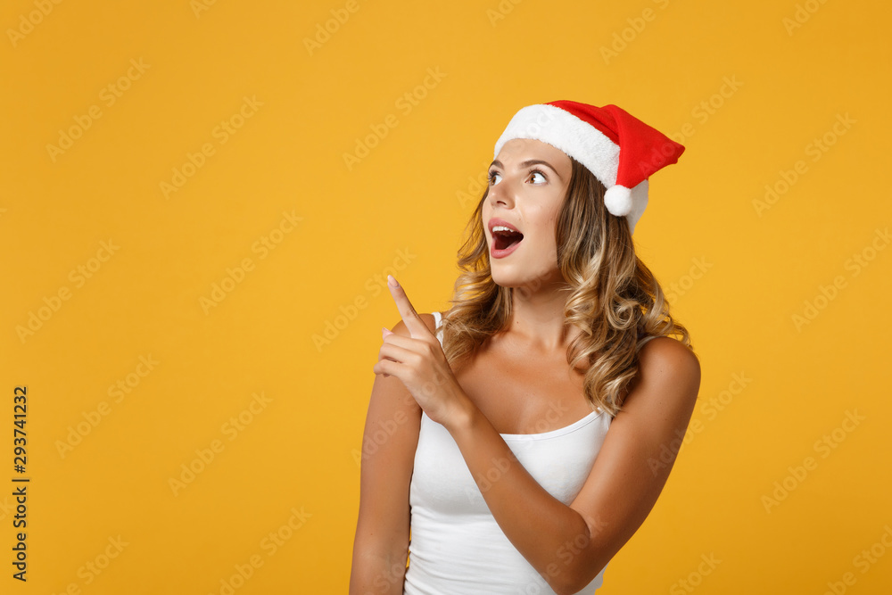 Shocked young Santa girl in white shirt, Christmas hat posing isolated on yellow background in studio. Happy New Year 2020 celebration holiday concept. Mock up copy space. Pointing index finger aside. Shocked young Santa girl in white shirt, Christmas hat posing isolated on yellow background in studio. Happy New Year 2020 celebration holiday concept. Mock up copy space. Pointing index finger aside.