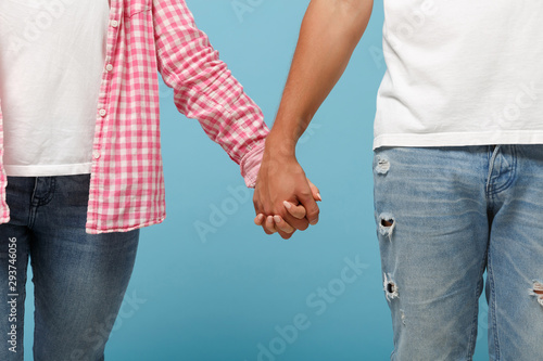 Cropped image of couple two friends guy girl in white pink empty blank design t-shirts posing isolated on pastel blue background in studio. People lifestyle concept. Mock up copy space. Holding hands.